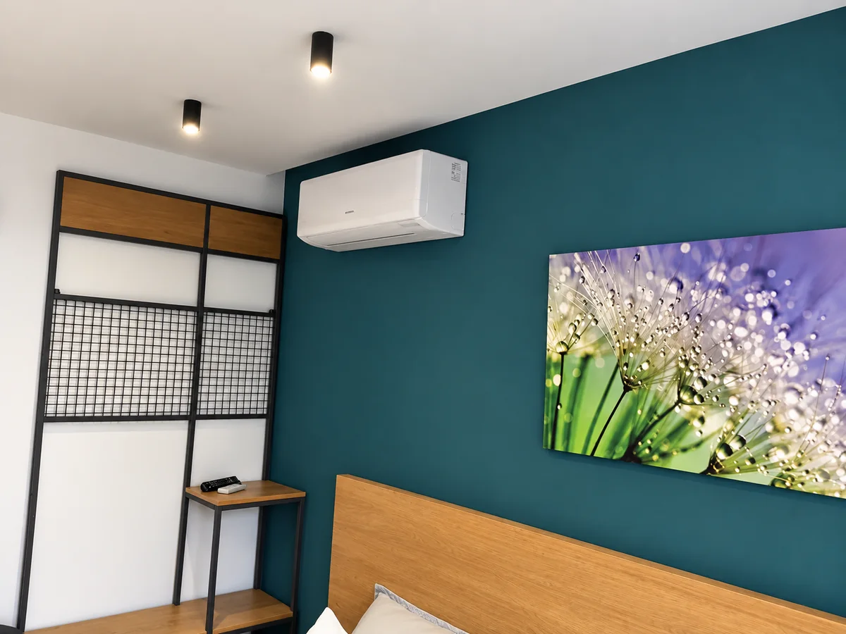 Wall-mounted cooling unit above a bed against a blue-green wall