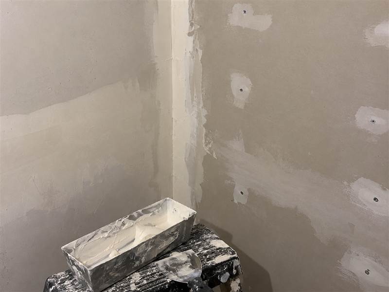 Unfinished drywall corner with compound patches and a pan on a work stool